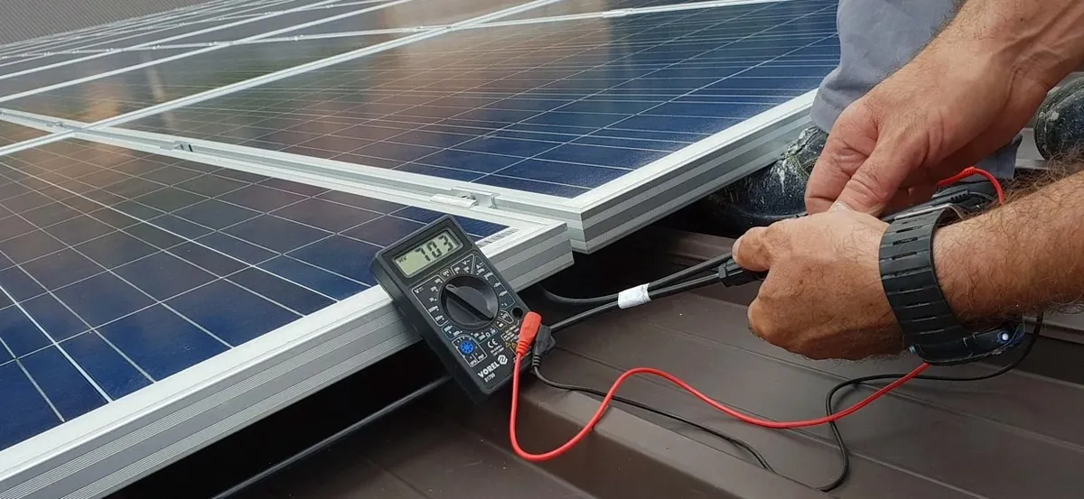 Hands holding a multimeter probe against the frame of a solar panel on a roof (illustrative photo)