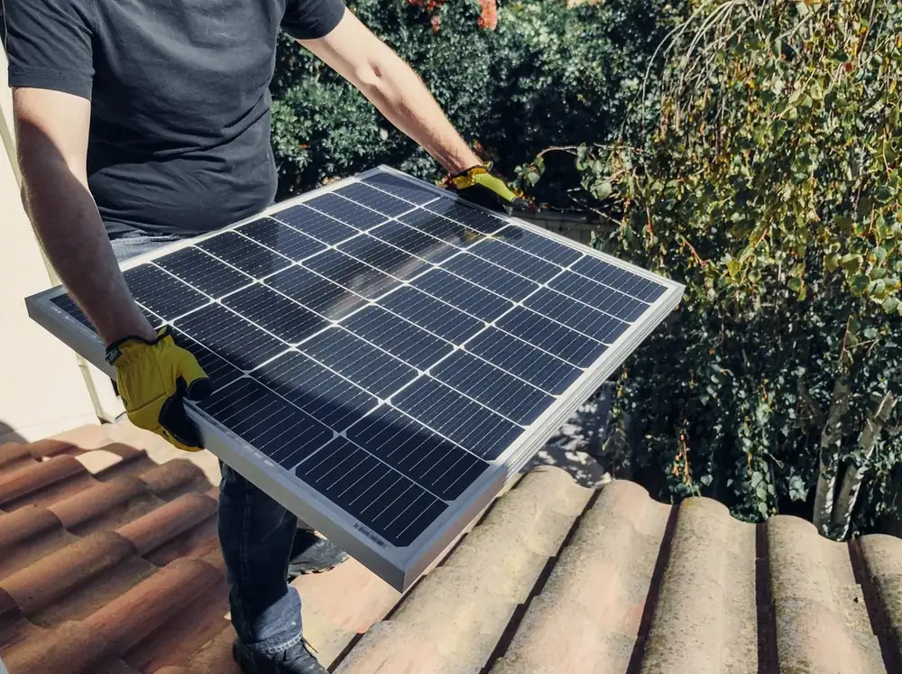 Person carrying a solar panel across a tiled roof (illustrative photo)